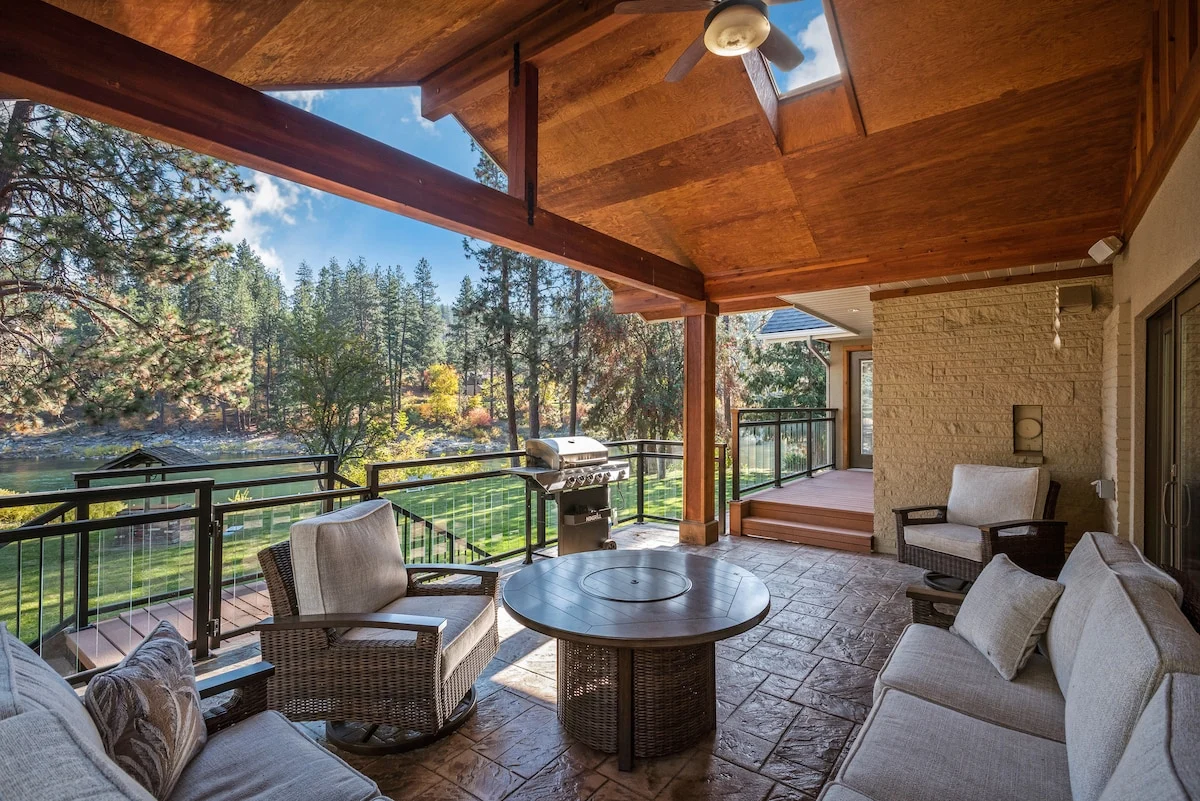 The River House covered patio with timber beams, outdoor seating, grill, and views over the rolling lawn to the Spokane River