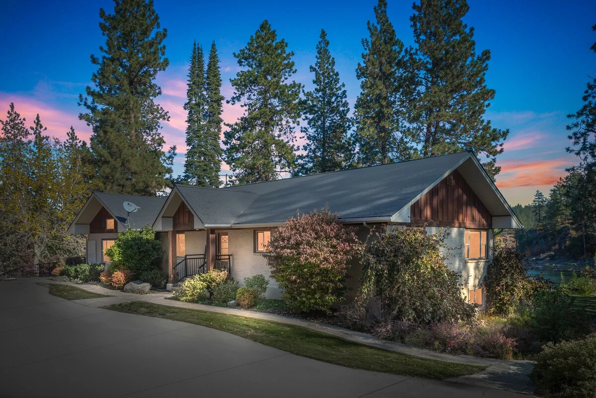 The River House exterior at dusk — home framed by tall pines with the Spokane River visible at the edge of the property